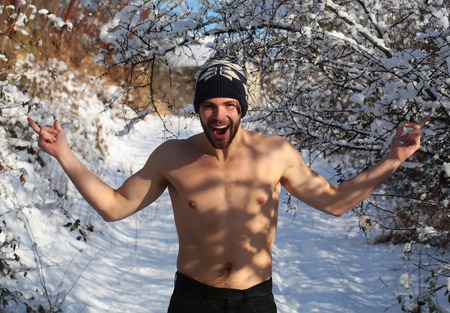 happy handsome bearded young man or sexy muscular guy with bare torso and chest on fit body in winter hat sunny outdoor in trees with snow on natural backgroundの写真素材