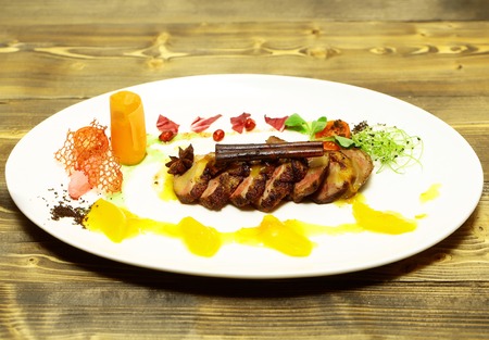 Roasted meat slices with cinnamon stick, spices, vegetables and yellow fruit jelly sauce on white plate on wooden background. Modern molecular gastronomyの写真素材