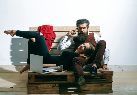 Young couple spend time together with laptop, computer. Pretty girl or beautiful woman sleeps on knees of handsome man, bearded hipster with beard, talking on mobile phone on wooden pallet sofaの写真素材