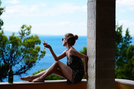 Cute female holding glass with white wine in hand with open bottle sitting on railing of balcony and looking at sea on natural background.の写真素材