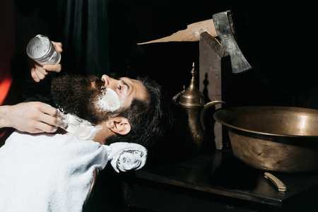 Handsome bearded man, hipster, brunette with beard and moustache has shave in barbershop. Barber works with vintage razor, shaving brush and foamの写真素材