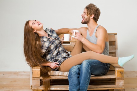 Young happy couple of pretty girl or cute woman, with brunette long hair and handsome man or muscular macho with muscular torso, sits on wooden with tea cups, pallet sofa on grey backgroundの写真素材