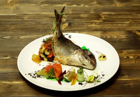 Roasted white fish served with colorful vegetables, zucchini, green jelly caviar, carrot, lime, sprouts on plate on wooden background. Modern molecular gastronomyの写真素材