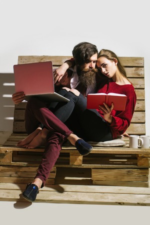 Young couple spend time together on wooden pallet sofa. Handsome man, bearded hipster with beard uses laptop, computer. Pretty girl or beautiful woman reads bookの写真素材