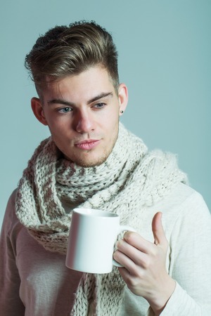Handsome sexy man portrait in white knitted winter scarf has fashionable hair and serious face. Young stylish guy with earring holds tea cup, drinking in studio on grey backgroundの写真素材
