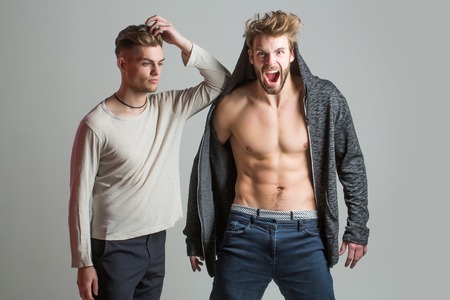 Handsome young bearded men friends in shirt and bare torso has fashionable hair and serious face on grey background. Stylish sexy guys with muscular body shouting in studioの写真素材