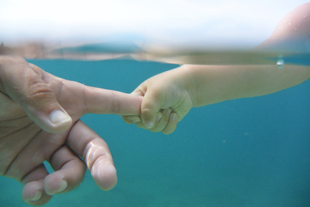 Baby hand holding dads finger in clear blue water in day.の写真素材