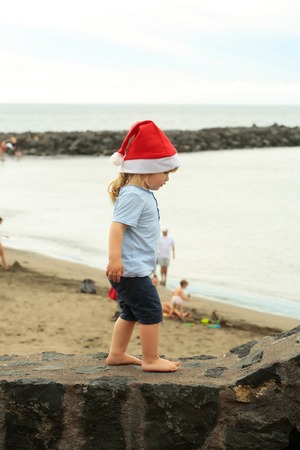 Cute baby boy in red santa hat with blond hair ponytail in blue tshirt and shorts walks barefoot on stone wall by sea beach on summer day on natural backgroundの写真素材