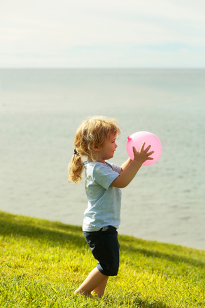 Cute baby boy with blond hair ponytail in blue tshirt and shorts plays with pink toy balloon on green grass by sea on sunny summer day on natural backgroundの写真素材