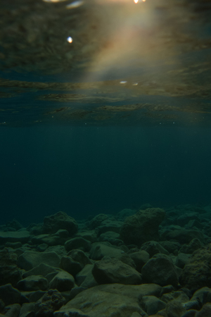 Underwater view of seabed with stones and gravel and sunbeam passing through water in evening.の写真素材