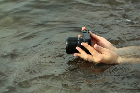 Female hands hold modern black digital camera or digicam over sea or ocean water surface outdoors on...