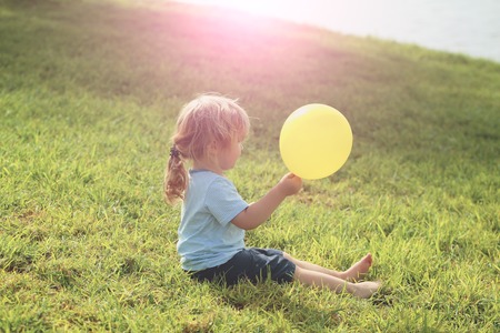 Cute baby boy with blond hair ponytail in blue tshirt and shorts sits on green grass with yellow toy balloon by sea in sunlight on sunny summer day on natural backgroundの写真素材