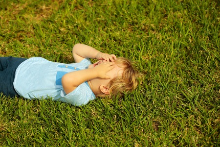 Crying cute baby boy with blond hair in blue tshirt hides face with hands lying on green grass outdoors on sunny summer day on natural backgroundの写真素材