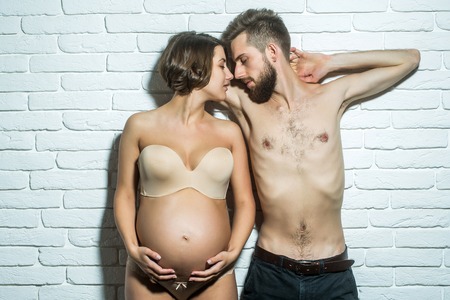 family parent couple of pretty happy sexy woman or cute pregnant girl with round belly in bra and handsome bearded man hipster with slim bare torso and chest on brick wall background at mothers dayの写真素材