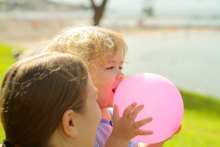 Mother and son, cute baby boy, with blond hair play with pink toy balloon on green grass by sea on sunny summer day on natural background at mothers dayの写真素材