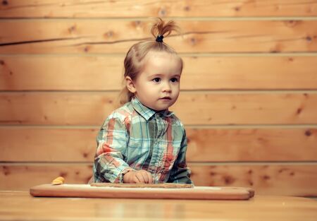 adorable small child chef or cute baby boy in fashionable chekered shirt cooking on board with flour, dough and knife on wooden backgroundの写真素材