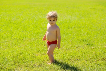 Cute baby boy with blond hair in red trunks sunbathes outdoors on sunny summer day on idyllic lawn on green grass backgroundの写真素材