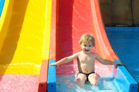Happy cute baby boy with blond hair rides from waterslide in water or aqua park outdoors on sunny...
