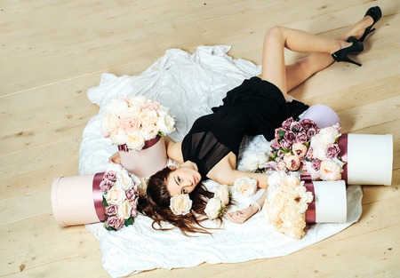 Sexy young pretty woman or cute girl with long hair in black dress and shoes laying among lilac rose and peony flowers on white bedsheet on wooden floorの写真素材