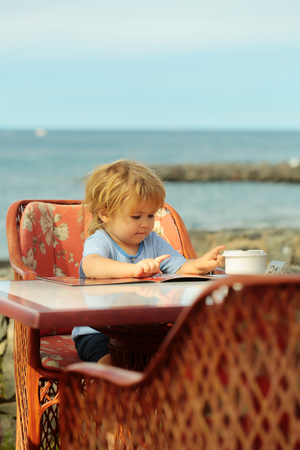 Cute baby boy with blond hair in blue tshirt reads menu card sitting at table in outdoor cafe by sea on sunny summer day on blurred natural backgroundの写真素材