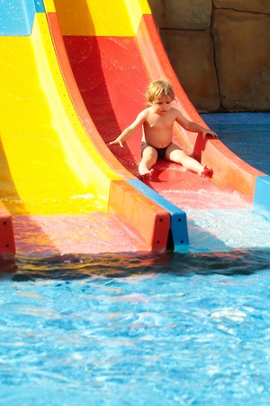 Cute baby boy with blond hair rides from waterslide in water or aqua park outdoors on sunny summer day on colorful backgroundの写真素材