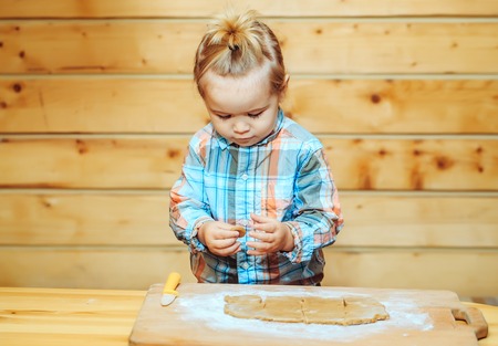 adorable small child chef or cute baby boy in fashionable chekered shirt cooking on board with flour, dough and knife on wooden backgroundの写真素材