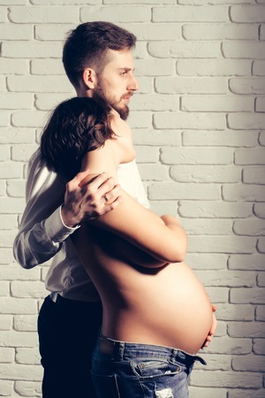 family parent couple of pretty smiling sexy woman or cute pregnant girl with naked round belly or abdomen and handsome bearded man hipster in white shirt on brick wall background at mothers dayの写真素材