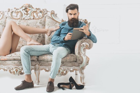 Handsome bearded man in blue shirt and denim with book and bare female legs touch him on luxurious baroque couch on white background, copy spaceの写真素材