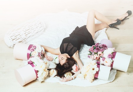 Sexy young pretty woman or cute girl with long hair in black dress and shoes laying among lilac rose and peony flowers on white bedsheet on wooden floorの写真素材