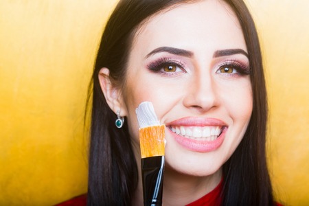 sexy cute woman smiling face or pretty girl with brunette long hair and lipstick on lips has fashionable makeup on yellow background, holds artist brushの写真素材