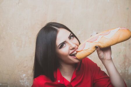 sexy cute woman or pretty girl with brunette long hair and makeup on happy smiling face in fashionable red shirt holds eats big burger or sandwich with sausage and cheese on textured wall backgroundの写真素材