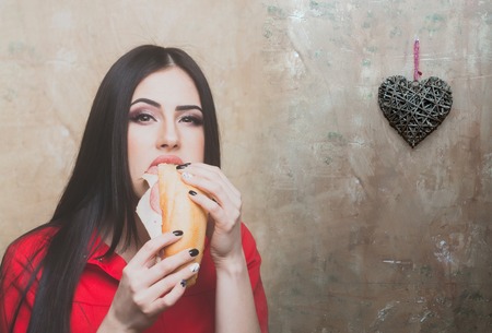 sexy cute woman or pretty girl with brunette long hair in fashionable red shirt holds eats big burger or sandwich with sausage and cheese on textured wall background with valentines heartの写真素材