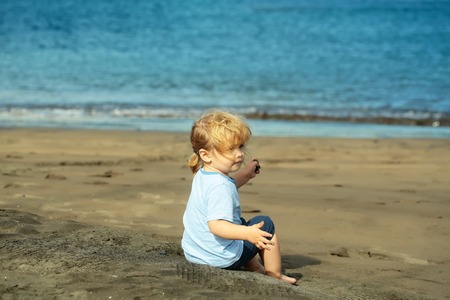 Cute baby boy with blond hair ponytail in blue tshirt and shorts sits on sand on sandy beach outdoors on sunny, summer day on blue sea backgroundの写真素材