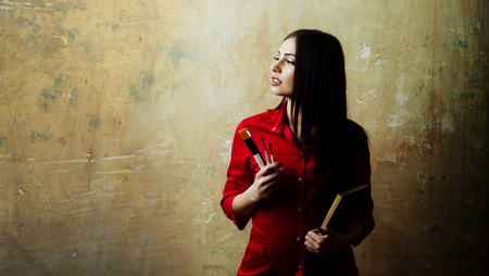 sexy cute woman or pretty girl with brunette long hair and makeup on face in fashionable red shirt holds brush set and notebook on textured wall background, copy spaceの写真素材