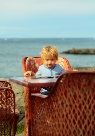 Cute baby boy with blond hair in blue tshirt reads menu card sitting at table in outdoor cafe by sea on sunny summer day on blurred natural backgroundの写真素材