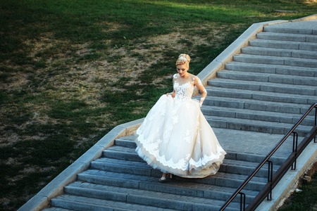 Pretty girl or cute bride with beautiful makeup and blond hair, hairstyle, in long white sexy wedding dress walking down grey stairs on sunny summer day outdoors on green grass backgroundの写真素材