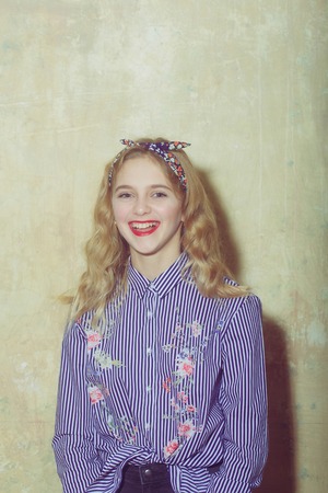 Cheerful pretty girl or beautiful woman with red lips and blond hair in blue striped shirt and stylish headband smiles indoors on beige wall backgroundの写真素材