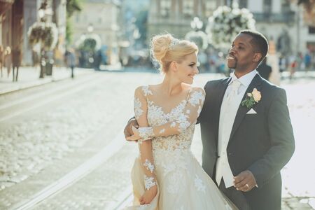 Handsome man or african American groom hugging pretty girl or cute bride with beautiful blond hair in white sexy wedding dress outdoors on blurred city street backgroundの写真素材