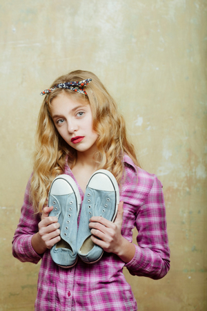 Pretty girl or beautiful woman with red lips and blond curly hair in stylish headband and violet plaid shirt with blue fashion sneakers in hands indoors on beige wall backgroundの写真素材