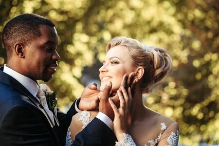 Loving groom or african American man gently touches face of pretty girl or adorable bride with beautiful make up and blond hair sunny summer day outdoors on blurred natural backgroundの写真素材