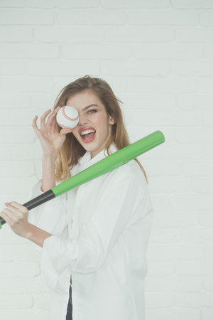 young pretty woman or cute sexy girl with beautiful long hair and red lips on adorable smiling happy face in fashionable white shirt holds green baseball bat or racket and ball on brick wall backgroundの写真素材