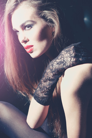 Pretty girl or sexy woman with beautiful makeup and long brunette hair wearing black lace gloves sits in light on dark backgroundの写真素材