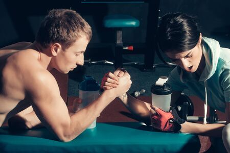 arm wrestling couple of pretty woman or cute girl and handsome guy or macho man with muscular torso and sexy body training in sport gym at trainer of weight lifting with water cupsの写真素材