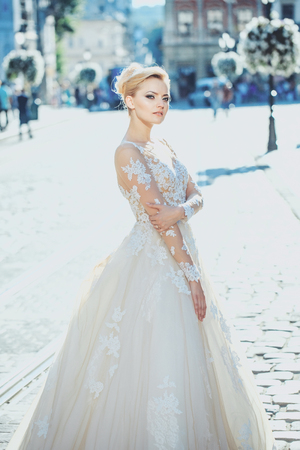 Pretty girl or cute bride with beautiful makeup and blond hair, hairstyle, in white sexy wedding dress poses on sunny summer day outdoors on blurred city street backgroundの写真素材