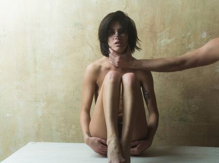 young pretty woman or sexy girl with naked chest and male hand touch body on textured wall background sitting on tableの写真素材