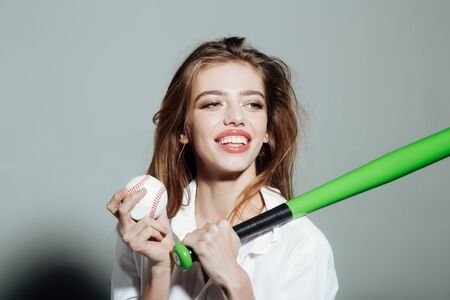 young pretty woman or cute sexy girl with beautiful long hair and red lips on adorable smiling happy face in fashionable white shirt holds green baseball bat or racket and ball on grey backgroundの写真素材