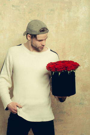 bearded macho man, brutal caucasian hipster with moustache in sweater and cap holds red rose flowers in box as valentines day present on textured wall backgroundの写真素材