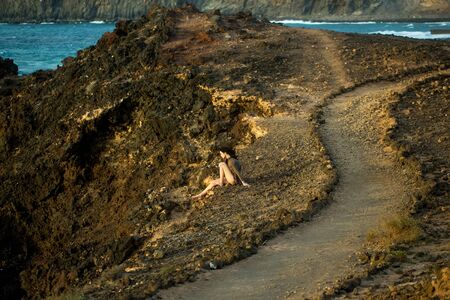 Pretty girl or beautiful woman, sexy slim brunette, sitting on mountain slope, brown rocky surface, on sunny summer day outdoors on blue sea backgroundの写真素材