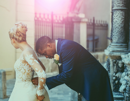 Handsome man or african American groom buttons white sexy wedding dress for pretty girl or cute bride with beautiful blond hair, hairstyle, outdoors in ancient town. Vintage filter colorの写真素材