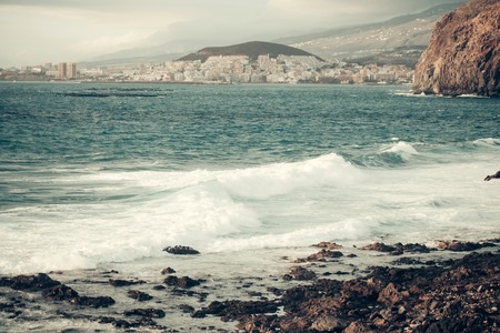 Beautiful blue sea or ocean water with stormy waves crashing against rocky coast with idyllic resort town on mountain backgroundの写真素材